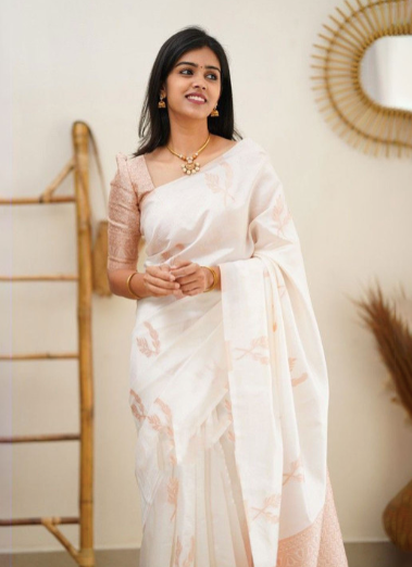Woman in a white saree with pink floral patterns standing indoors.