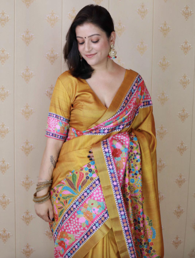 Woman wearing a yellow saree with colorful patterns against a patterned wall.