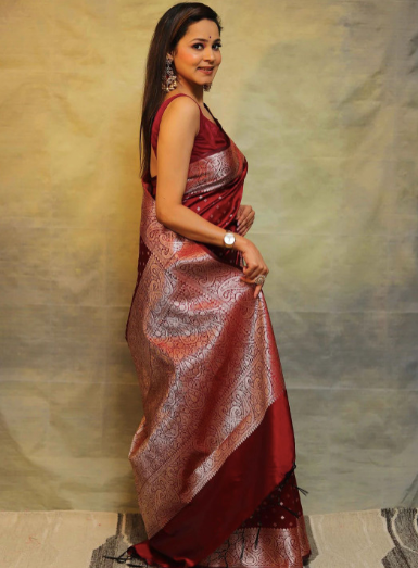 Woman wearing a red saree with a gold border against a textured wall.