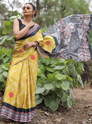 Woman in a yellow saree with a floral blouse standing outdoors among greenery.