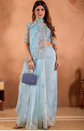 Woman in a light blue traditional outfit with a purple handbag against a beige background