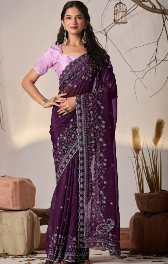 Woman wearing a purple saree with a white blouse, standing indoors with decorative elements.