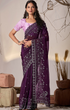 Woman wearing a purple saree with a white blouse, standing indoors with decorative elements.