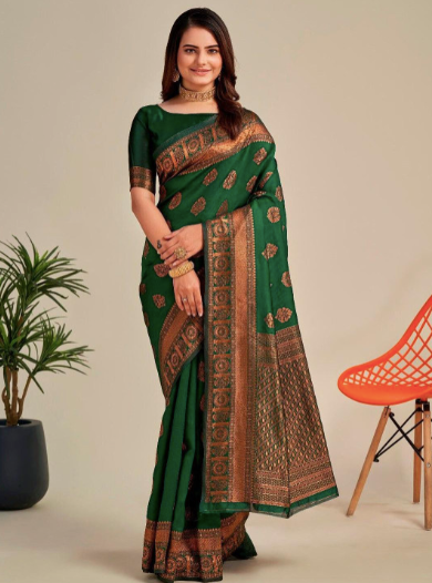Woman wearing a green saree with a patterned border, standing next to an orange chair and a plant.