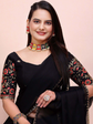 Woman wearing a black saree with floral embroidery and colorful jewelry against a pink background