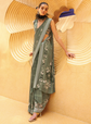 Woman in a green saree with floral patterns against a yellow textured wall.