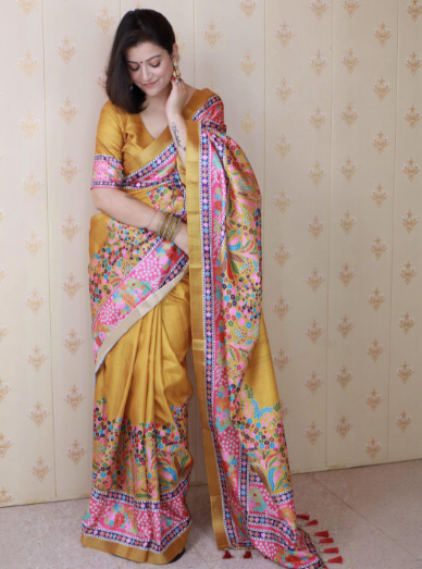 Woman wearing a yellow saree with colorful patterns against a beige wall.