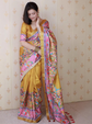 Woman wearing a yellow saree with colorful patterns against a beige wall.
