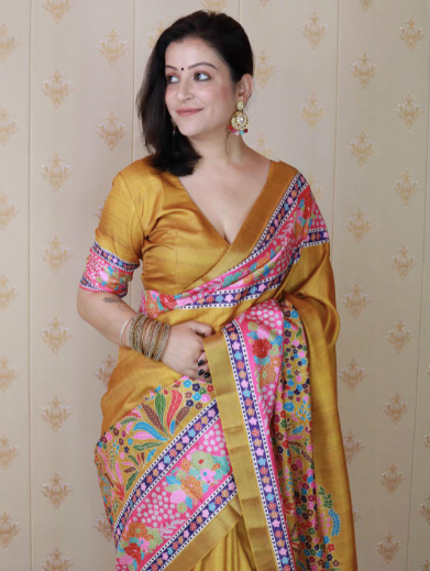 Woman wearing a yellow saree with colorful patterns against a beige wall.