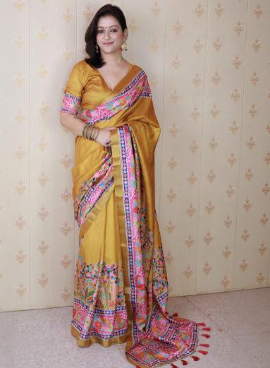 Woman wearing a yellow saree with a colorful border against a patterned wall.