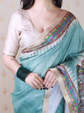 Woman wearing a turquoise saree with colorful border, holding it with one hand.