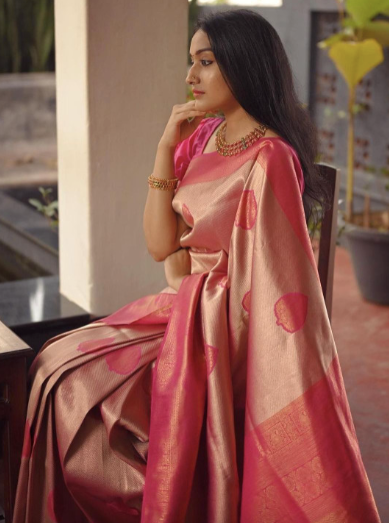 Woman in a pink saree with a gold border sitting outdoors.