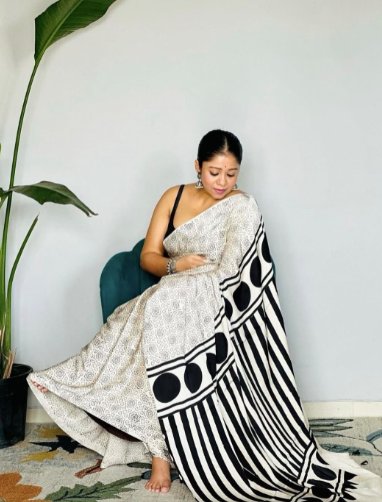 Woman sitting on a chair with a white and black patterned fabric draped over her lap, against a light blue wall.
