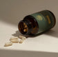 Brown glass bottle with a green label and gold emblem, containing capsules on a beige surface.