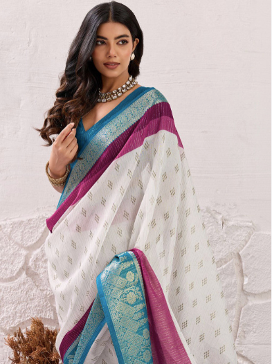 Woman wearing a white saree with blue and purple accents against a white brick wall.
