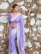 Woman wearing a purple saree with a white border against a stone wall.