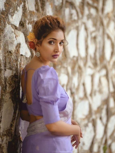 Woman in a purple saree leaning against a textured wall.